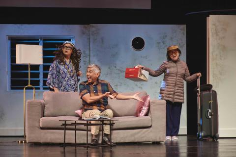 Image of Siti Khalijah in a curly long hair wig. Rody Vera is seated on the couch and Liow Shi Suen is standing next to it with a huge suitcase in hand.